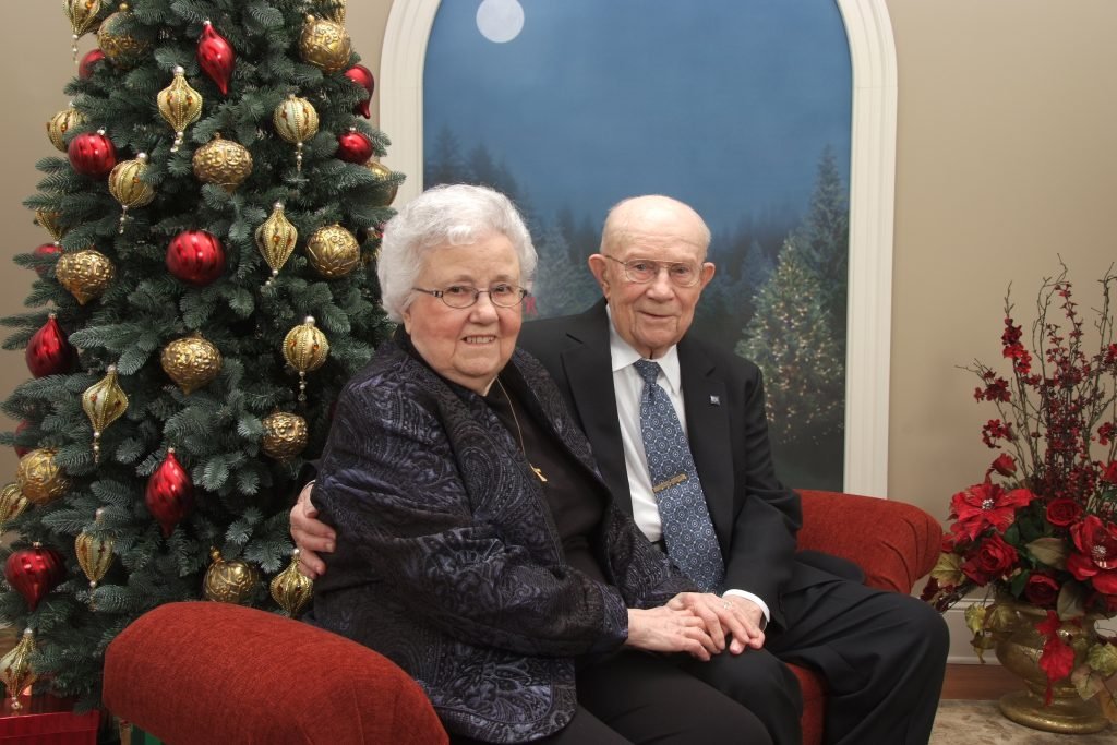 A lovely Christmas Photo of an elder couple sitting on a red bench in front of a Christmas tree.